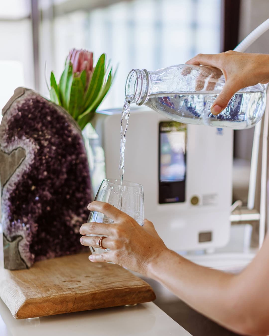 Alkaline ionized hydrogen-rich water being poured from a glass bottle beside an Enagic K8 ionizer on a kitchen counter